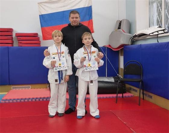 Тренер Андрей Абашин с двумя мальчиками в белых кимоно с дипломами под флагом России